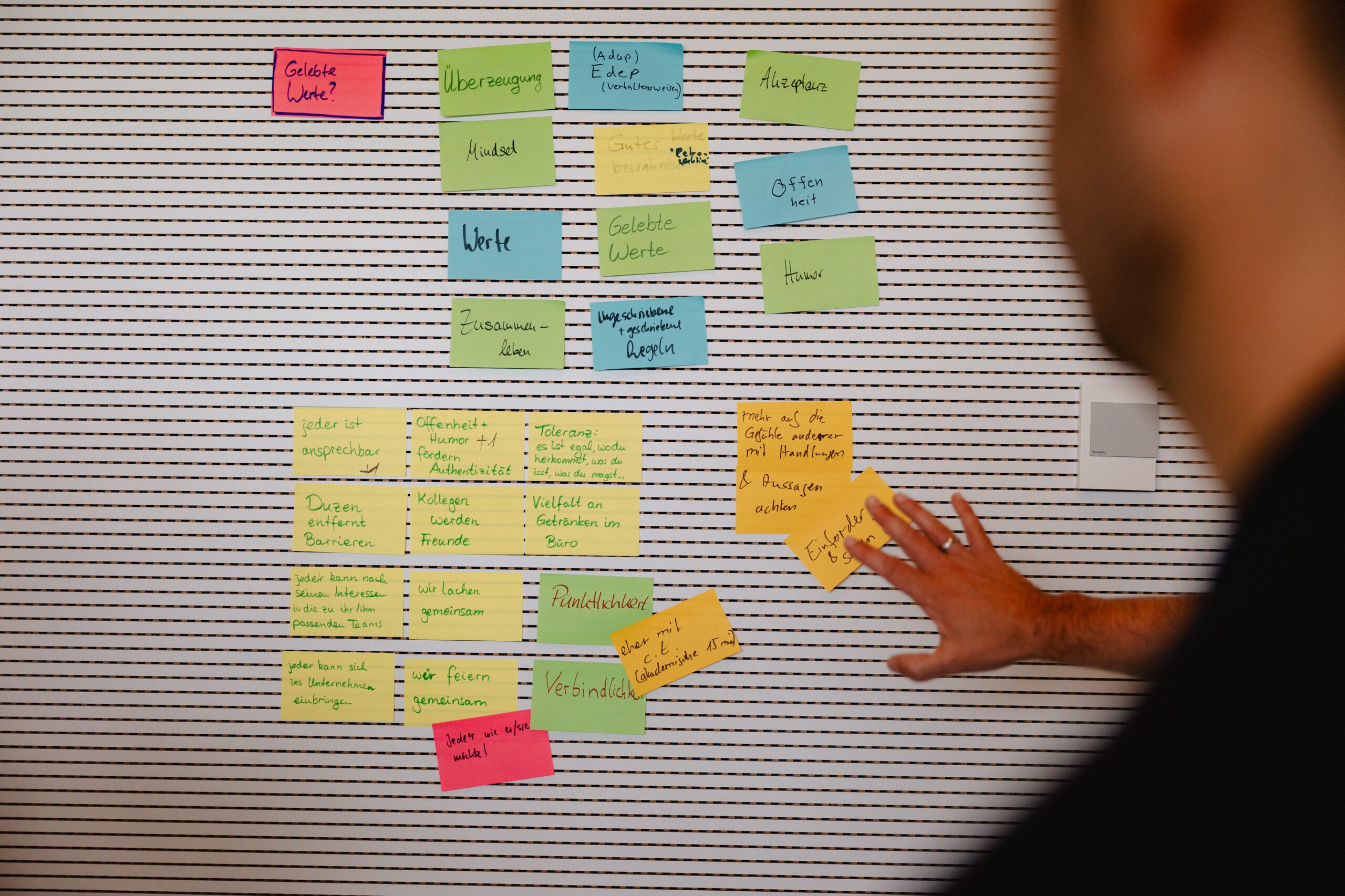 Im Vordergrund sieht man verschwommen einen Mann, der ein Sticky Note an eine Wand klebt.